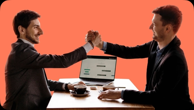 Dois homens em um ambiente de trabalho, vestindo ternos, sentados frente a frente em uma mesa com duas xícaras de café e um laptop ao fundo. Eles estão sorrindo e apertando as mãos. 